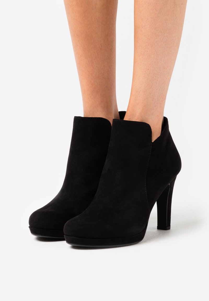 Bottines en daim noir à talons blocs hauts portées sur des jambes nues sur fond blanc.