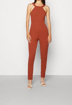 Kvinde iført en ærmeløs rustfarvet halter-jumpsuit med tilspidsede ben og hvide, remmede hælsandaler.