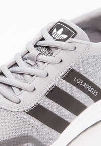 Grå idrottssko med mesh-övre, platta snören, tre svarta ränder och texten "LOS ANGELES". Gummisula och vadderad plös.