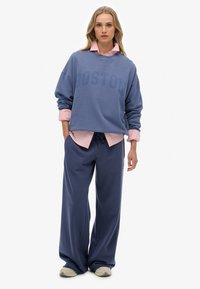 Sudadera de gran tamaño color azul marino con la impresión "BOSTON", sobrepuesta a una camisa de botones rosa claro, combinada con pantalones de chándal holgados en azul marino.