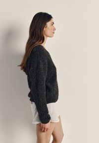 Sieviete ar gariem brūniem matiem, kas valkā tumšu pūkainu džemperi un gaisā pelēkas šortus, stāv profilā vienkārša bēša fona priekšā.