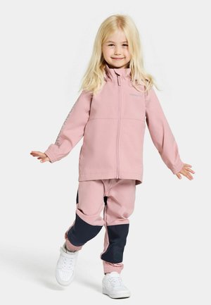 Veste coupe-vent rose avec un col montant et fermeture éclair, associée à un pantalon rose assorti avec des accents bleu marine. Chaussures de sport blanches.