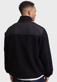 Homme portant une veste en polaire noire avec un col haut et un panneau en nylon sur l'épaule, tourné le dos contre un fond uni clair.