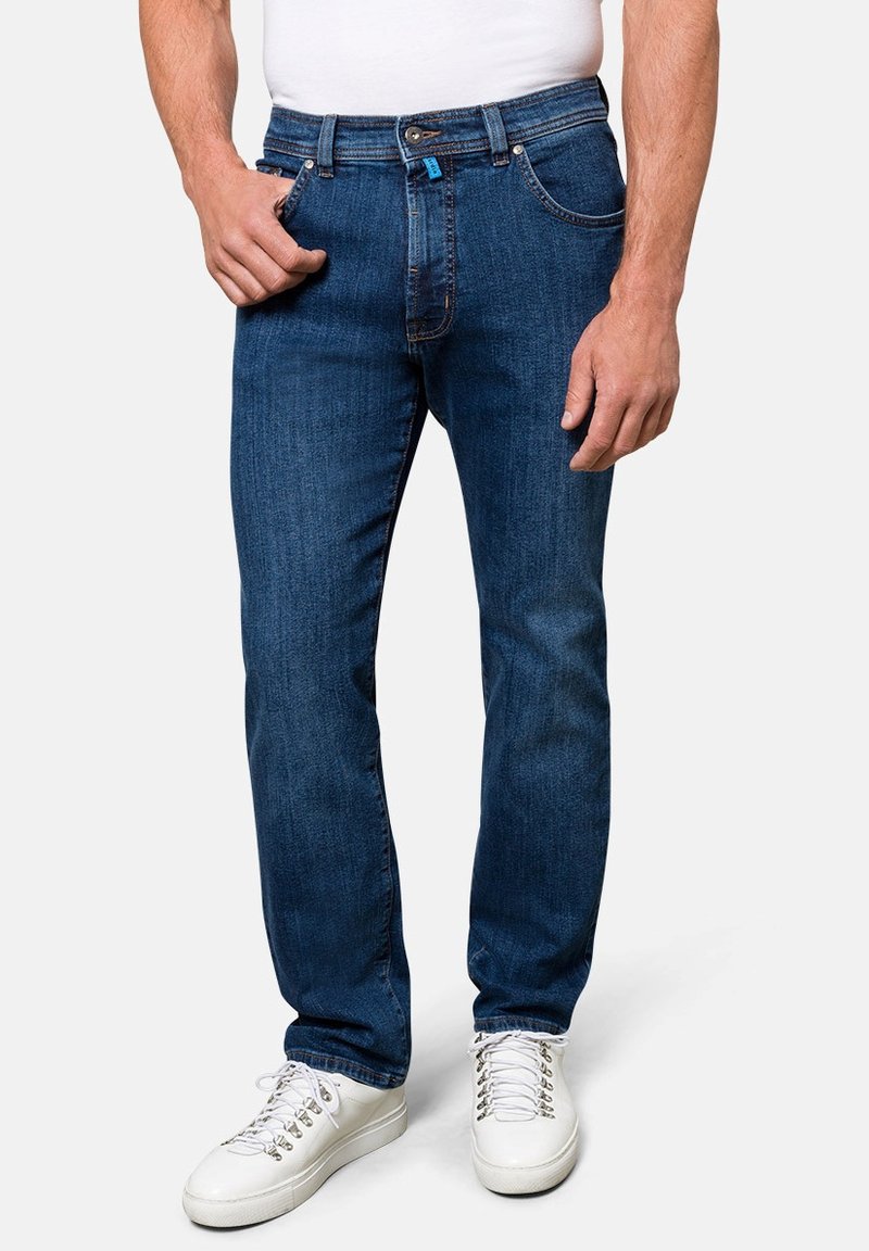Dunkelblaue Jeans aus Denim mit geradem Schnitt, fünf Taschen und Knopfverschluss, kombiniert mit weißen Sneakers und einem schlichten weißen Hemd.