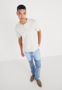 Krémovo sfarbená pruhovaná košeľa s okrúhlym výstrihom a tromi gombíkmi, spojená s modrými patchwork džínsami a hnedými semišovými čižmami.