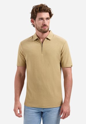 Man met kort krullend bruin haar en baard, die een beige poloshirt met korte mouwen en een ritssluiting aan de voorkant draagt en lichtblauwe spijkerbroek.