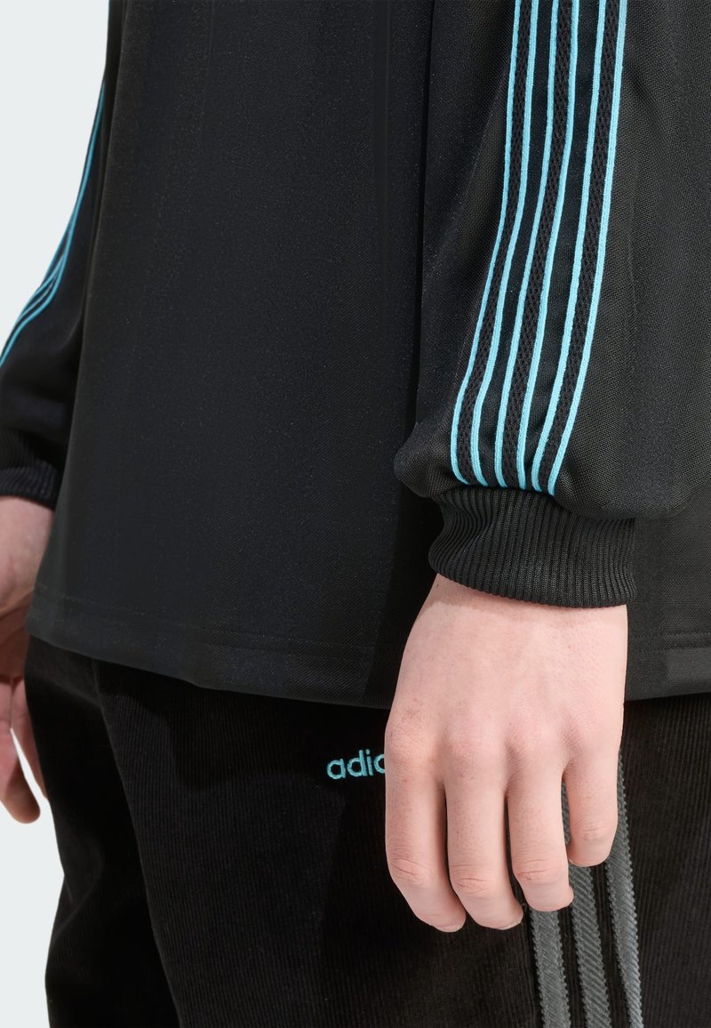 Zwart sportief shirt met lange mouwen, voorzien van drie lichtblauwe strepen op de mouwen, gedragen met zwarte corduroy broek waarop een adidas-logo zichtbaar is.