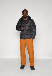 Casaco acolchoado preto com costuras visíveis e capuz, combinado com calças laranja. O conjunto é completado com calçado preto. Fundo liso.