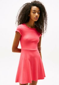 Korallenrosa Kurzarmkleid mit ausgestellt Rock, elegantem Wellen-Ausschnitt und weichem, strukturiertem Stoff. Kleines Logo-Accents auf der Brust.