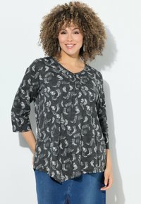 Mujer con cabello rizado, vistiendo pendientes de plata, una blusa oscura con estampado floral y mangas de 3/4, y jeans azules, sonriendo frente a una pared blanca.