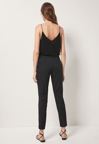 Vrouw met lang bruin haar, gekleed in een zwarte mouwloze top, zwart aansluitende broek en zwarte sandalen met banden, staande tegen een simpele lichte muur.