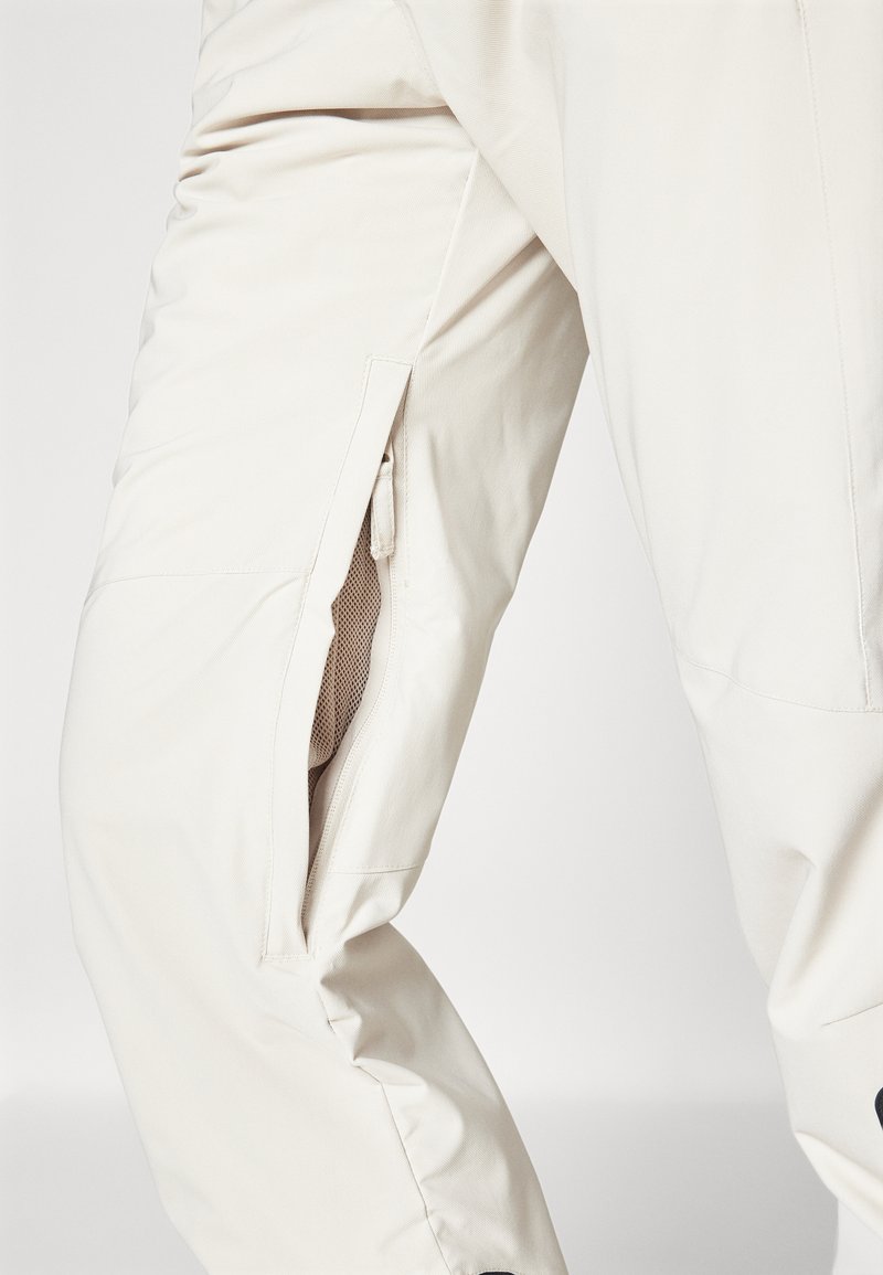 Beżowe, lekkie spodnie o teksturowanej powierzchni, z bocznymi kieszeniami na zamek i zwężającym się krojem nogawek.