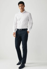 Bílá košile s dlouhými rukávy, kombinovaná s tmavými formálními kalhotami a černými společenskými botami. Model stojí na neutrálním pozadí.