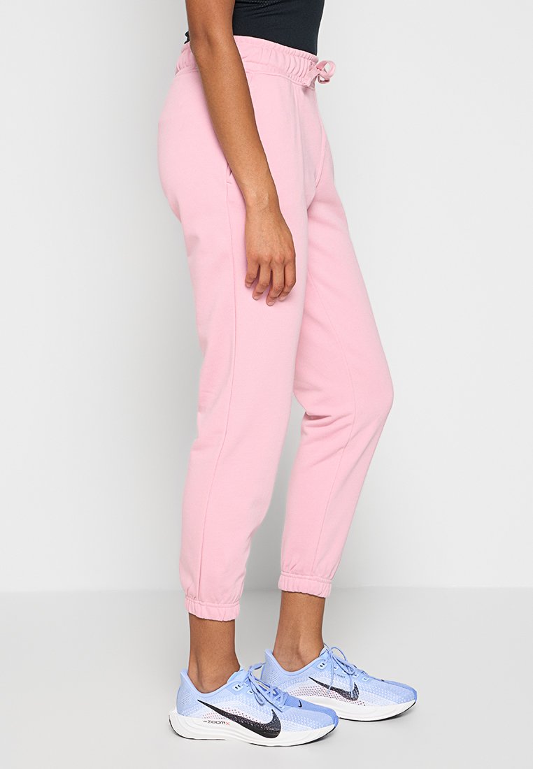 Osoba w jasnoróżowych dresach z elastycznymi mankietami i pasem, w połączeniu z jasnoniebieskimi butami do biegania Nike i czarną bluzką.
