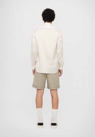 Camisa de Rayas blancas de manga larga con puños ajustados, combinada con pantalones cortos en beis claro y calcetines blancos. Zapatillas slip-on con detalles en negro completan el look.