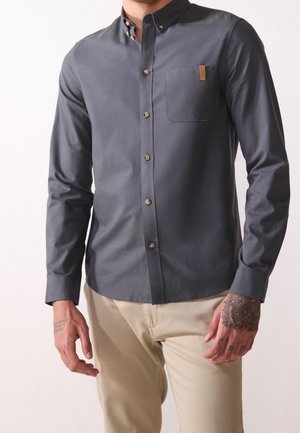 Mężczyzna ubrany w ciemnoszarą koszulę zapinaną na guziki z kieszenią na piersi i jasnobeżowe spodnie, pokazujący zadrukowane tatuażami dłonie na jednolitym tle.