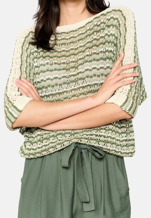 Femme croisant les bras, portant un pull tricoté rayé vert et crème et un pantalon vert olive avec une ceinture nouée à la taille.