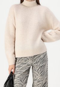 Femme portant un pull à col roulé beige et un pantalon imprimé zèbre gris, tenant un sac en cuir noir sur fond blanc.