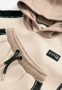 Beige hættetrøje med sort kant ved halsen, udstyret med snoretræk og et mærketag. Glat tekstur med et afslappet design og ribbet sømdetalje.