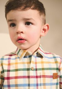 Chemise à carreaux avec des couleurs pastel : bleu, jaune, vert et rouge. Comprend une poche poitrine avec une étiquette brune et des boutons en bois.