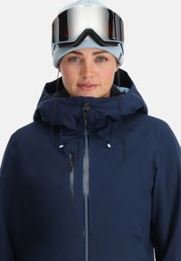 Frau trägt eine dunkelblaue Winterjacke mit hohem Kragen, eine hellblaue Strickmütze und reflektierende Skibrillen, die auf ihrer Stirn ruhen.