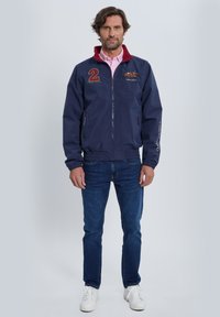 Chaqueta cortavientos azul marino con cuello rojo, que cuenta con cierre de cremallera, dos bolsillos frontales, acentos bordados y puños elásticos.