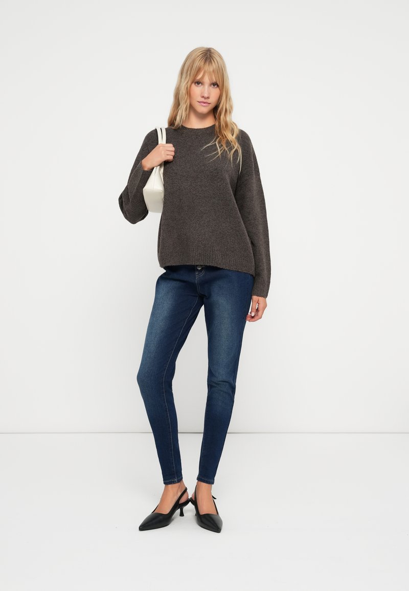 Pull en tricot gris foncé avec des manches larges, associé à un jean bleu foncé ajusté et des escarpins noirs à bout pointu. Le modèle porte un sac à main blanc.