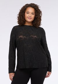 Schwarzes Langarmoberteil mit einem strukturierten floralen Spitzenmuster, rundem Halsausschnitt und lässiger Passform, kombiniert mit schwarzen Leggings.