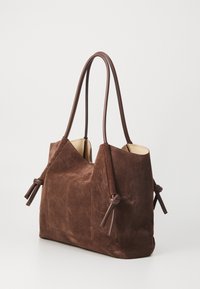 Sac fourre-tout en daim marron avec de longues poignées en cuir brun, présentant des accents noués sur les bords et un design simple à ouverture supérieure.