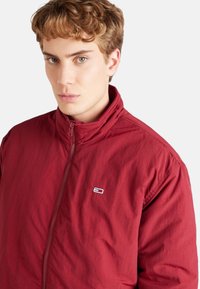 Chaqueta roja con cremallera y cuello alto. Fabricada en un tejido ligero que presenta un pequeño logo y detalles de costura visibles. Textura suave.