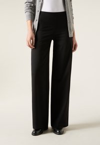 Pantalons larges noirs en tissu lisse avec une taille élastique. Portés avec un cardigan gris et des chaussures noires, offrant une coupe décontractée.