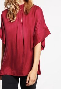 Burgundy silkes blus med hög hals, korta vida ärmar och en nyckelhålsdetalj framtill. Slät textur med en subtil lyster.