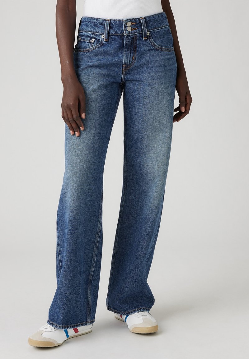 Personne portant un jean bleu taille haute, délavé moyen, à jambes larges, et des baskets blanches avec des rayures rouges et bleues.