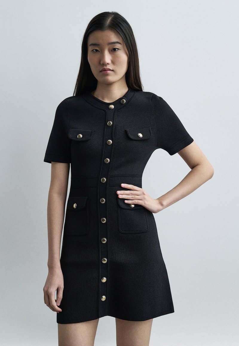 Νεαρή γυναίκα με μακριά σκούρα μαλλιά που φορά μ ια μαύρη κοντομάνικη φόρεμα με χρυσά κουμπιά και τέσσερις μπροστινές τσέπες με καπάκι, στέκεται με το ένα χέρι στη μέση.