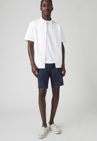 Cămașă albă cu mâneci scurte și nasturi, purtată peste un tricou alb, asortată cu pantaloni scurți bleumarin cu un model punctat. Adidași de culoare deschisă.