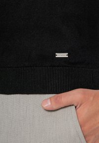 Czarny sweter z dzianiny z prążkowanym wykończeniem i małą srebrną metką WITTCHEN. Ręka umieszczona w kieszeni jasnoszarych spodni.