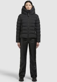 Sort puffjakke med høj krave, lynlås foran og sidelommer. Båret sammen med sorte læderbukser og støvler, præsenteret mod en neutral baggrund.