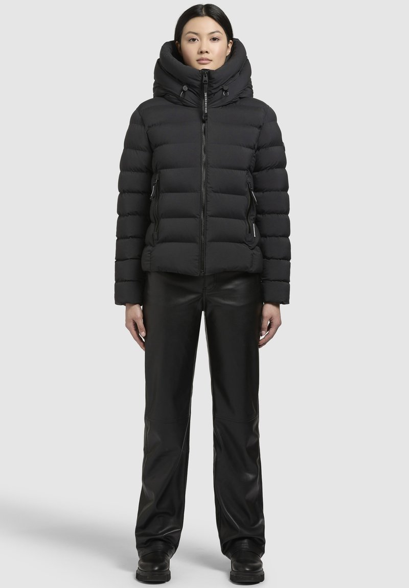 Sort puffjakke med høj krave, lynlås foran og sidelommer. Båret sammen med sorte læderbukser og støvler, præsenteret mod en neutral baggrund.