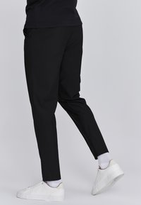 Zwarte broek met een taps toelopende pasvorm, gemaakt van gladde stof. Draagt witte sneakers en geribbelde sokken. Eenvoudig ontwerp, zonder opvallende patronen.