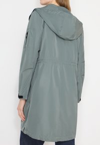 Personne portant un long imperméable à capuche bleu gris clair avec des poches latérales et une poche zippée sur la manche gauche, de dos.