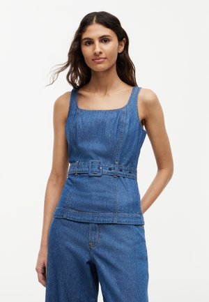 Kobieta ubrana w bezrękawnik z kwadratowym dekoltem z denimu, z dopasowanym paskiem w talii i luźnymi spodniami z denimu, stojąca na jednolitym tle.