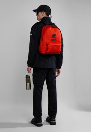 Sac à dos rouge avec une poche avant, porté par une personne en veste et pantalon noirs, tenant une bouteille d'eau en métal verte. Fond gris.