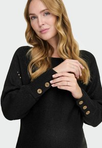 Maglione nero a maglia con scollatura ampia e texture a coste. Gli accenti a bottone sui polsini aggiungono dettagli al design.