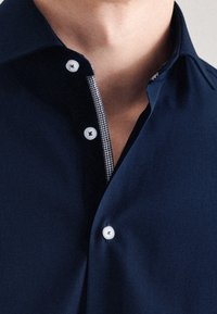 Camicia blu navy con colletto button-down, caratterizzata da bottoni bianchi e un motivo houndstooth all'interno del colletto e della patta. Texture liscia.