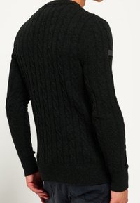Maglione a coste verde scuro con un motivo testurizzato. Presenta polsini e orlo a costine, e ha un piccolo logo sul manica.