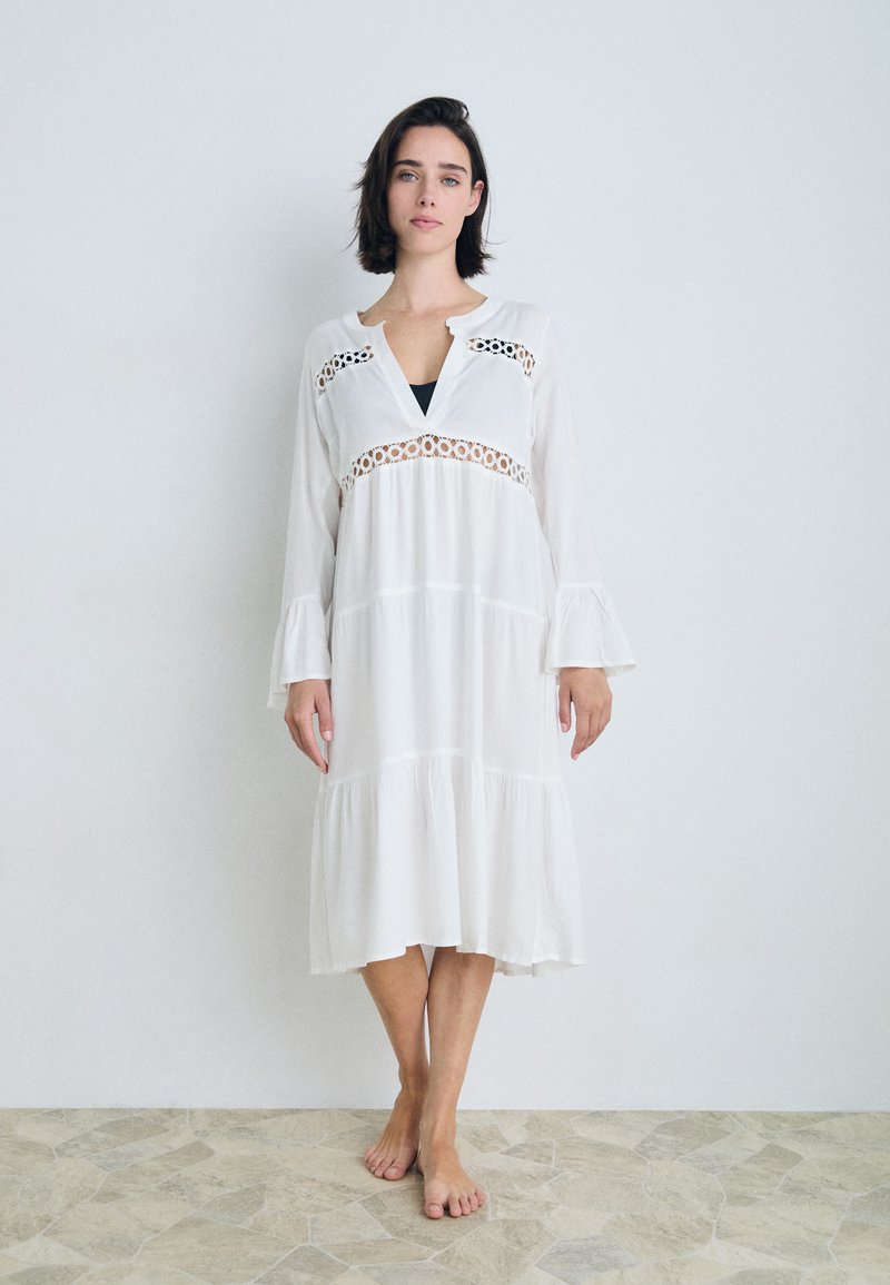Vestido longo branco com mangas com babados, detalhes em renda ao redor do decote e barra em camadas. Tecidos suaves, corte solto, usado descalço.