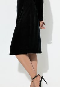 Czarna sukienka do kolan z miękkiego aksamitu, z długimi rękawami i dopasowanym krojem. Czarne szpilki z błyszczącymi paskami wokół kostki.