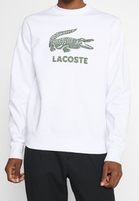 Sudadera blanca con el logotipo de Lacoste en patrón de reptil verde y texto debajo. Puños y dobladillo acanalados. Textura de tela suave.