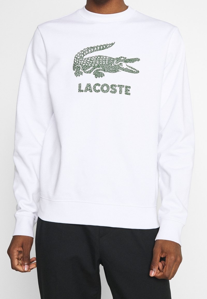 Sudadera blanca con el logotipo de Lacoste en patrón de reptil verde y texto debajo. Puños y dobladillo acanalados. Textura de tela suave.