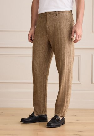 Mann, der braune strukturierte Hose und schwarze Loafers trägt, steht auf Holzfußboden vor cremefarbenen Wandpaneelen.